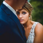 A bride in a lace wedding dress leans against a man in a navy suit, looking directly at the camera, with greenery in the blurred background.