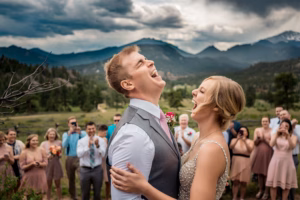 A bride and groom laugh together outdoors during their wedding ceremony, surrounded by applauding guests with mountains and a cloudy sky in the background.
