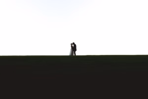 A couple dressed in wedding attire stands embracing on a grassy hilltop against a plain white sky.