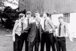 A group of six young men in dress shirts and ties, with one in a suit, stand outside by a wooden building, laughing and gesturing casually.