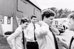 Four young men in dress shirts and ties stand outside near a barn, talking and adjusting their clothes. A car and trees are visible in the background. The image is in black and white.