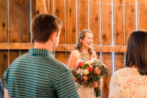 A woman holding a large bouquet of flowers smiles while standing in front of two people indoors, with wooden walls in the background.