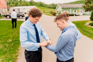 Two men in blue shirts and dark ties stand on a paved path; one helps the other with his cufflinks while two more people are visible in the background.
