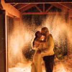 A couple in formal attire kisses under a wooden structure at night, with snow falling and illuminated in the background, creating a dramatic effect.