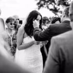 A bride in a strapless dress wipes her eye while holding paper, surrounded by guests at an outdoor wedding ceremony.