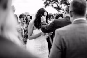A bride in a strapless dress wipes her eye while holding paper, surrounded by guests at an outdoor wedding ceremony.