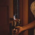 A person in formal attire is partially visible through a doorway, while another person in a white garment stands in the foreground holding the door handle.