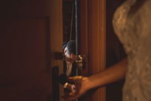 A person in formal attire is partially visible through a doorway, while another person in a white garment stands in the foreground holding the door handle.