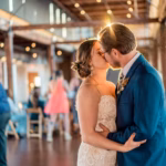 A bride and groom kiss while dancing together at their wedding reception in a warmly lit venue with guests in the background.