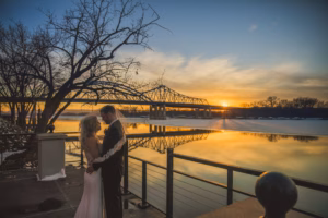 A couple in wedding attire stands close together by a riverside at sunset, with a bridge and bare trees in the background.