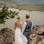 A bride and groom stand on rocky terrain overlooking a lake, with mountains and snow-capped peaks in the background.