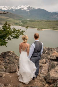 A bride and groom stand on rocky terrain overlooking a lake, with mountains and snow-capped peaks in the background.