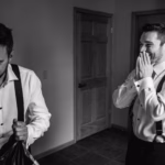 Two men in dress shirts and suspenders stand indoors; one is smiling with hands over his mouth while the other looks down, holding a bag.