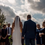 A bride and groom stand holding hands, facing a group of people outdoors under a partly cloudy sky.