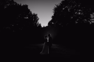 Silhouetted couple standing close together outdoors at dusk, framed by trees, with a faint light illuminating their outlines.