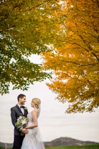 A bride and groom stand outdoors, facing each other and smiling, with green and yellow autumn trees and a distant hill in the background.