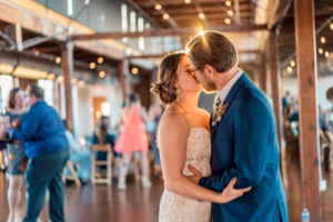 A bride and groom share a kiss while dancing at their wedding reception in a warmly lit venue with guests in the background.