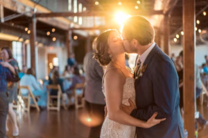 A bride and groom kiss on the dance floor of a warmly lit wedding reception venue, with guests seated in the background and sunlight shining behind them.
