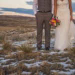 A bride and groom stand side by side outdoors on partially snowy ground, with the bride holding a colorful bouquet. The photo is cropped at their shoulders.