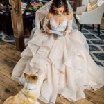A bride in a white wedding dress sits indoors reading a note, while a small dog looks up at her on a wooden floor.