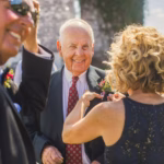 A woman pins a boutonniere onto an older man's lapel outdoors, while another man in sunglasses smiles nearby.