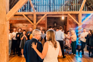 A couple dances together in the center of a wooden barn while a group of people stand in the background, watching and socializing.