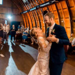 A bride and groom dance together in a warmly lit barn while guests watch and smile in the background.