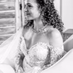 A woman in a lace off-shoulder wedding dress and veil sits, smiling, with her hair styled in loose curls and wearing a necklace.
