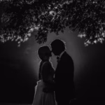 A couple stands close together under a tree at night, silhouetted against a bright light in the background.