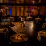 A bride and groom share a first dance in the center of a dimly lit, elegantly decorated reception hall with round tables and chandeliers.