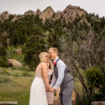 A bride and groom hold hands and smile at each other outdoors, with rocky hills and green trees in the background.