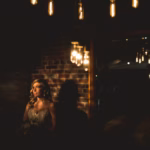 A woman in a wedding dress stands in a dimly lit room with exposed brick walls and hanging light bulbs.