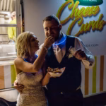 A bride in a white dress feeds a groom in a vest and tie outside a food truck labeled "Cheese Curds" at night.