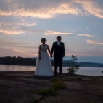 A couple in formal wedding attire holds hands and walks beside a lake at sunset, with trees and clouds in the background.