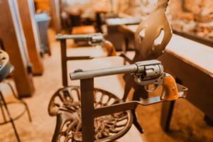 Close-up of two chairs designed with revolver-shaped armrests, positioned in a rustic indoor setting with blurred background details.