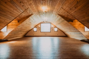 Spacious, empty attic with wooden floors and sloped wood-paneled ceiling, illuminated by natural light from a central window and two smaller side windows.