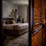 Two people are in a dimly lit bedroom; one is sitting on the bed while the other stands behind, appearing to do their hair or makeup. The room has brick walls and framed pictures.