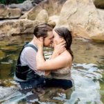 A couple in formal attire sits waist-deep in a clear rocky river, facing each other closely and touching foreheads with hands on each other's faces.