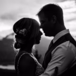 A couple stands close together in silhouette, facing each other outdoors with mountains and a setting sun in the background.