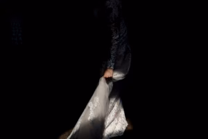 A person in a lace dress is partially illuminated against a dark background, holding the lower part of the dress with one hand.
