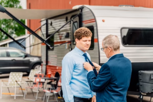 Two men in suits stand outside near a camper trailer; one adjusts the other's tie. Folded chairs and a parked car are visible in the background.