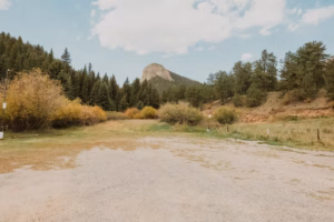 A gravel clearing bordered by grassy fields, trees, and hills, with a rocky mountain peak in the background under a partly cloudy sky.