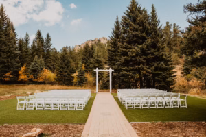 Rows of white chairs face a white wedding arch outdoors, surrounded by tall evergreen trees and a mountain landscape under a partly cloudy sky.
