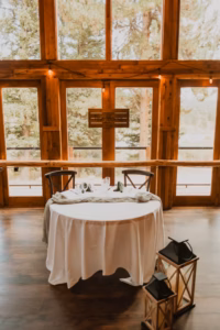 A round table with a white tablecloth is set for two in front of large wooden windows; two lanterns sit on the floor beside the table.