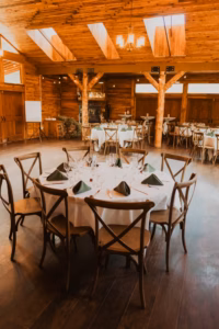 Round table with white tablecloth, set with glassware and black napkins, surrounded by wooden chairs in a rustic wooden banquet hall with large windows and high ceilings.