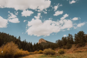 A rocky hill rises above a forested landscape under a partly cloudy sky, with grassy fields and autumn foliage in the foreground.
