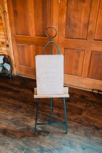 A sign on an easel stands on a wooden floor in front of a wooden door. The text on the sign is partially illegible.