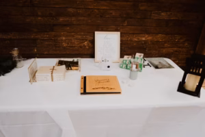 A white table with a guestbook, framed sign, instant camera, photo props, and a lantern, set up against a wooden wall.