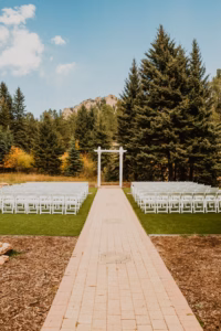 Outdoor wedding ceremony setup with rows of white chairs facing a white arch, surrounded by trees and mountains in the background.