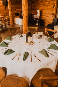A round table set for six with green napkins, multiple wine glasses, wooden chairs, and a lantern centerpiece in a rustic wooden venue.